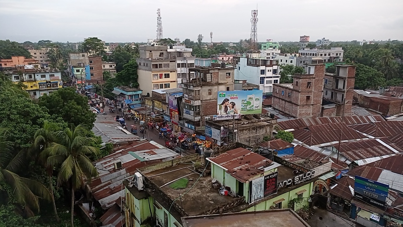 A streetscape in Lakshmipur, with small and large buildings and a mix of flat and sloped roofs.